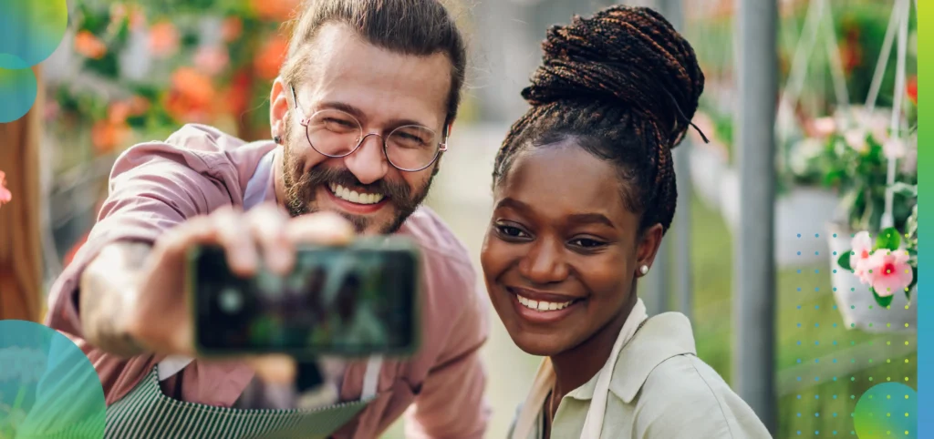 Multiracial couple of gardeners working in a greenhouse and taking selfie with a smartphone.