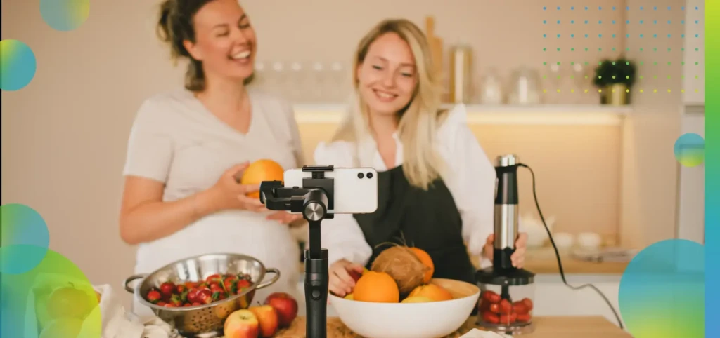 Two happy millennial woman bloggers recording video of cooking smoothie using smartphone in the kitchen.